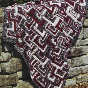 May include: A handmade crochet blanket with a geometric pattern in burgundy, white, and gray. The blanket is draped over a stone wall, showcasing its intricate design and textured surface. The color palette creates a cozy and inviting feel.