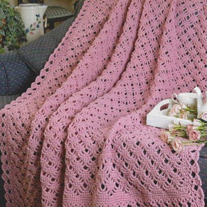 Puede incluir: Una manta de ganchillo rosa empolvado con un diseño calado detallado y un borde festoneado, sobre un sillón estampado. Una bandeja blanca con una taza y flores descansa sobre el brazo de la silla.