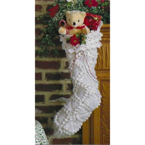 May include: White crocheted Christmas stocking with a teddy bear and red poinsettia inside. The stocking is hanging on a wooden mantel.