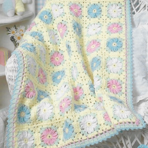 May include: A crocheted baby blanket with a daisy pattern. The blanket has a yellow base with squares of white, pink, and blue daisies. It is trimmed with a light blue and pink scalloped edge. The blanket is draped over a white wicker chair.