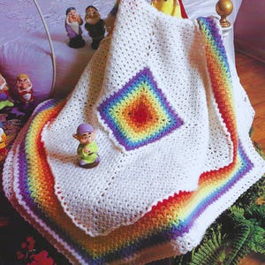 May include: A white crocheted baby blanket with a rainbow border and a rainbow square in the center. The blanket is draped over a bed with a purple headboard and a white bedspread.