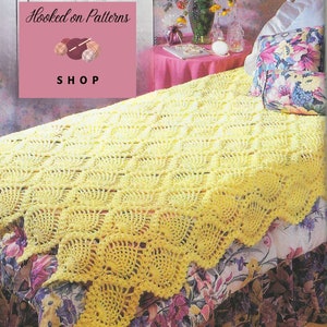 Pode incluir: Uma manta de crochê amarela com uma borda festonada, exibida em uma cama com uma colcha estampada de flores. A manta é feita com um padrão repetitivo de círculos. A imagem inclui uma placa rosa com o texto "Hooked on Patterns SHOP".