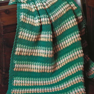 May include: A hand-crocheted blanket in shades of green, cream, and tan. The blanket has a textured pattern with horizontal stripes and a diagonal design. The blanket is draped over a wooden chest, showing its soft texture and colour variations.