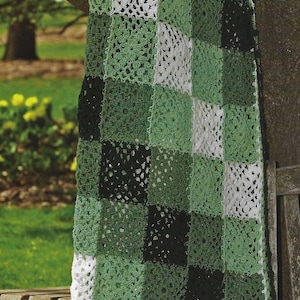 May include: A handmade crochet blanket in a patchwork design, featuring squares of white, various shades of green, and black. The blanket is draped over a wooden bench and tree branch, showcasing its texture and color variations.