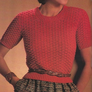 Könnte beinhalten: Ein roter Strickpullover mit kurzen Ärmeln und einem runden Ausschnitt. Der Pullover wird mit einem braunen Ledergürtel und einem gemusterten Rock getragen.