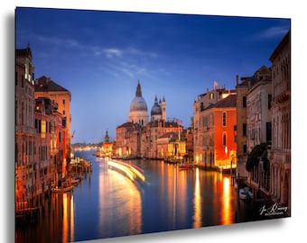 May include: A framed print of Venice at dusk. The image captures the Grand Canal with illuminated buildings lining the waterway. A long exposure shows the blurred lights of a boat moving through the water, under a deep blue sky.