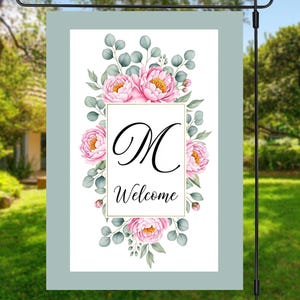 May include: A garden flag with a floral design. The flag features pink peonies, green leaves, and the letter "M" in elegant script, with the word "Welcome" below. The flag is framed in a light blue border and displayed on a black metal stand.