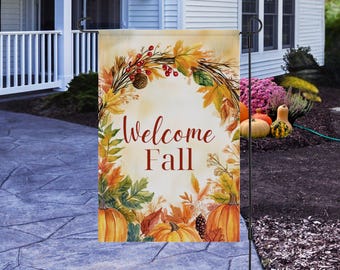 May include: A decorative garden flag with the words "Welcome Fall" in a script font. The flag features a wreath of fall leaves, berries, and pinecones, with pumpkins at the bottom. The background is a light beige.
