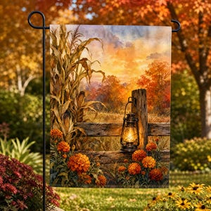 May include: A rectangular garden flag featuring a printed autumn scene with a wooden fence, stalks of corn, and bright orange marigolds. A lit metal lantern hangs from the fence post against a backdrop of a sunset sky and fall foliage.