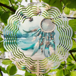 May include: A metal wind spinner with a blue and white dreamcatcher design. The dreamcatcher has feathers and beads. The wind spinner is in front of a green leafy background.