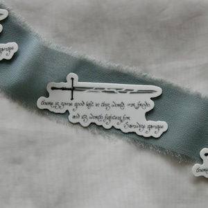 May include: Three white stickers with a sword design and text are arranged on a teal ribbon against a white background. The stickers have a black sword illustration and script. The ribbon has a frayed edge.