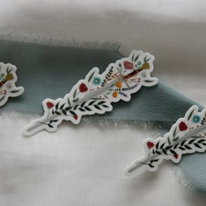 Puede incluir: Tres pegatinas decorativas con un diseño floral y de flecha. Las pegatinas tienen un fondo blanco con una ilustración detallada de una espada, flores y hojas. Las pegatinas están dispuestas sobre una tela blanca y azul.