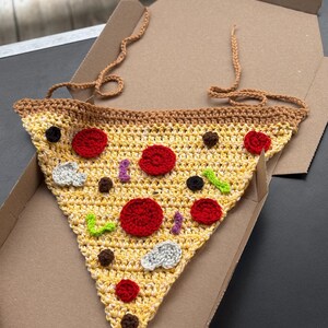 May include: A crocheted pizza slice with red, black, and white toppings, including pepperoni and mushrooms. The pizza is yellow and brown, with a brown crust and a tan strap. It is inside a cardboard pizza box.