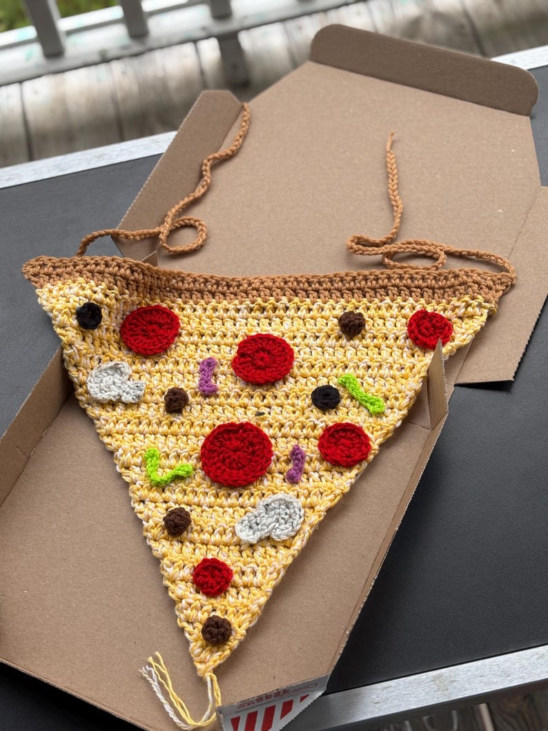 May include: A crocheted pizza slice with red, black, and white toppings, including pepperoni and mushrooms. The pizza is yellow and brown, with a brown crust and a tan strap. It is inside a cardboard pizza box.