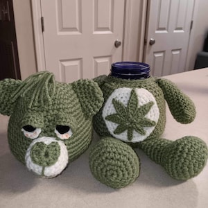 May include: A green crocheted bear-shaped jar cover with a white and green cannabis leaf design on the belly. The bear has a separate head, a small ball, and a blue glass jar inside.