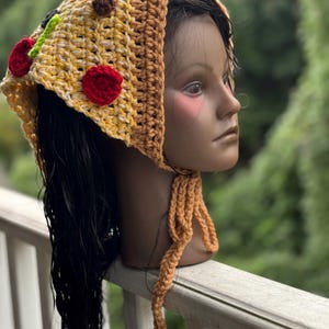 May include: A handmade crochet hat in the shape of a pizza slice. The hat features yellow, brown, red, and green yarn, with red and brown yarn embellishments to resemble toppings. The hat has long, braided ties.