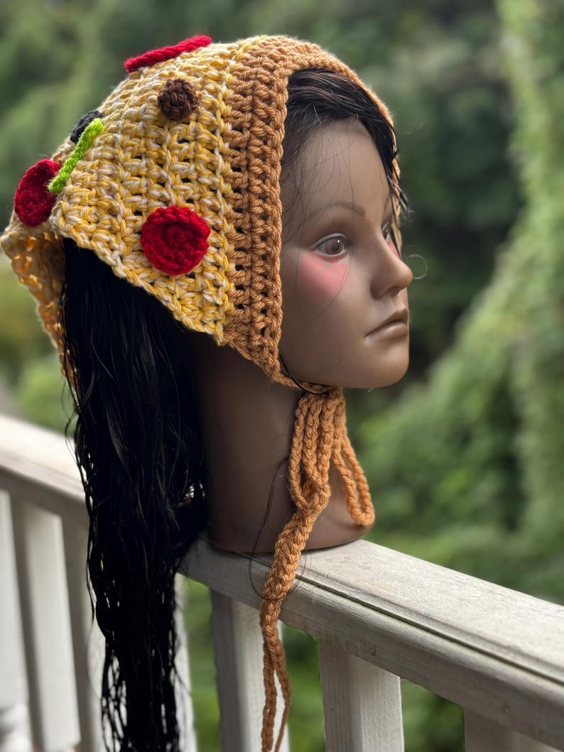 May include: A handmade crochet hat in the shape of a pizza slice. The hat features yellow, brown, red, and green yarn, with red and brown yarn embellishments to resemble toppings. The hat has long, braided ties.