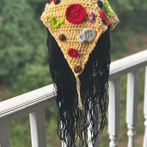 May include: A handmade, crocheted pizza hat with a yellow base, red pepperoni, and various colorful toppings. The hat is triangular in shape and has long, black hair attached, creating a unique and playful accessory.