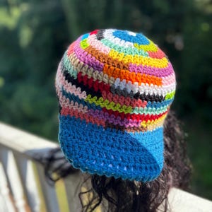 May include: A colorful, handmade crochet hat with a blue brim. The hat features a vibrant mix of colors including blue, pink, yellow, orange, and green. The hat is a unique, eye-catching accessory.