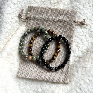 May include: Three beaded bracelets in green, brown, and black sit on a small beige drawstring bag. The bracelets are made of natural stone beads. The background is a soft, white, textured surface.