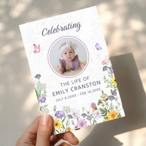 May include: A rectangular memorial card featuring the text Celebrating THE LIFE OF EMILY CRANSTON JULY 8, 20XX - FEB 14, 20XX. A circular photo of a baby wearing a white headband is centered, surrounded by watercolor illustrations of colorful wildflowers and butterflies.