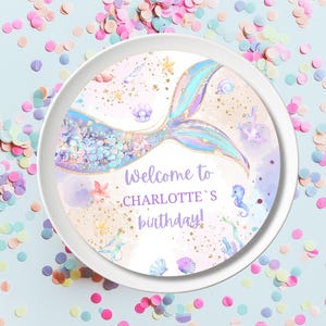 Puede incluir: Un plato de papel redondo blanco con un diseño de cola de sirena. La cola es iridiscente con purpurina dorada y escamas moradas, azules y rosas. El plato tiene un borde morado con el texto "¡Bienvenido al cumpleaños de Charlotte!"