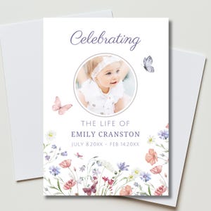May include: A white card with the text "Celebrating" in a script font. A photo of a baby is in a circular frame. The text "The Life of Emily Cranston" is below the photo, along with the dates July 8.20XX - Feb 14.20XX. Watercolor flowers and butterflies decorate the card.