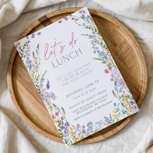 May include: A white invitation card with the text "let's do LUNCH" in pink script, surrounded by a floral border of purple, blue, and yellow flowers. The card is on a wooden plate.