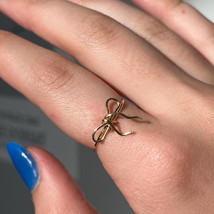 May include: A gold-colored ring with a bow design. The ring is on a finger with blue nail polish.
