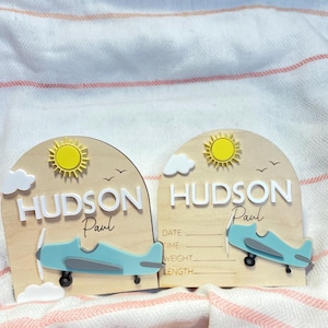 May include: Two wooden plaques with a light brown background, each featuring a blue airplane, a yellow sun, and the name "Hudson Paul" in white. One plaque includes lines for date, time, weight, and length. White clouds and birds are also present.