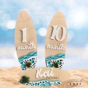 May include: Wooden surfboard-shaped monthly milestone markers with a blue and green ocean wave design. The markers are numbered 1 and 10. A smaller surfboard-shaped sign reads "Kai Michael".