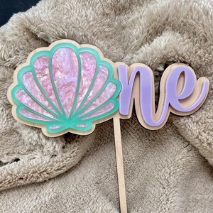 May include: A wooden cake topper with a seashell and the word "one". The seashell is pink and white with a teal outline. The word "one" is lavender with a wooden base. The topper is designed for a first birthday celebration.