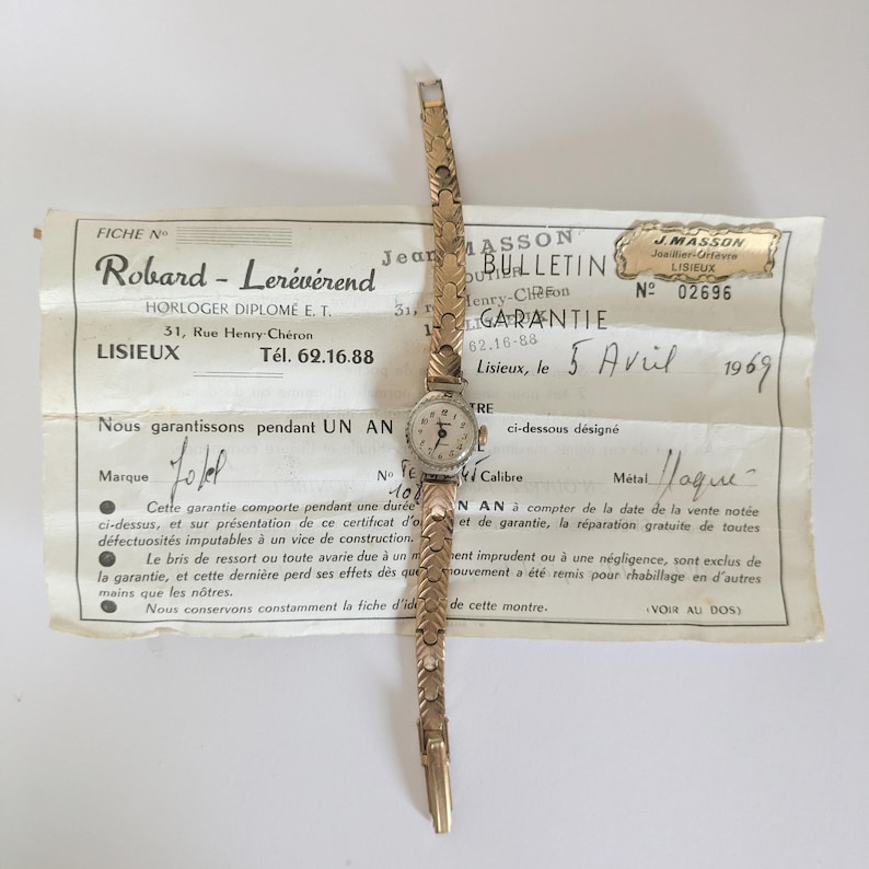 May include: A vintage gold-toned wristwatch with a small round face and a delicate, patterned band. The watch rests on a faded document with French text, including the date "5 Avril 1969". The watch is a classic timepiece.