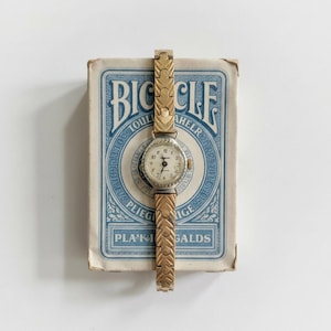 May include: A vintage gold-toned wristwatch with a round face and a light-colored dial. The watch is set on top of a blue and white Bicycle playing card box. The watch band has a decorative link design.
