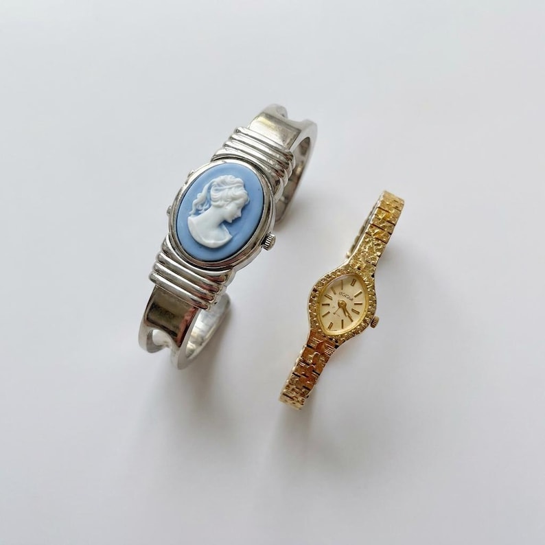 May include: Two vintage watches. One is a silver-tone cuff bracelet watch with a light blue cameo face. The other is a gold-tone watch with a textured band and a small oval face.