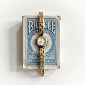 May include: A gold-toned wristwatch with a small, round white face and black hands. The watch band features a woven design with black and brown accents. The watch is displayed on a vintage Bicycle playing card box with blue and white text.