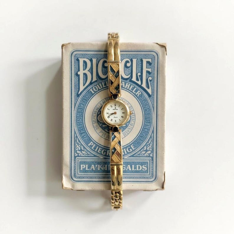 May include: A gold-toned wristwatch with a small, round white face and black hands. The watch band features a woven design with black and brown accents. The watch is displayed on a vintage Bicycle playing card box with blue and white text.