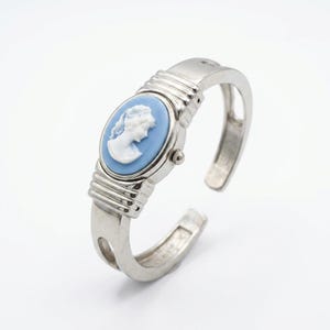 May include: Silver-toned cuff watch with an oval, light blue face featuring a white cameo profile. The watch has a classic design with a hinged opening and a ribbed band. A small dial is visible on the side of the watch face.