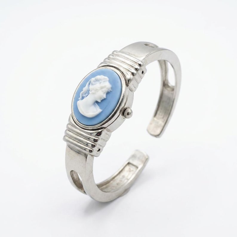 May include: Silver-toned cuff watch with an oval, light blue face featuring a white cameo profile. The watch has a classic design with a hinged opening and a ribbed band. A small dial is visible on the side of the watch face.