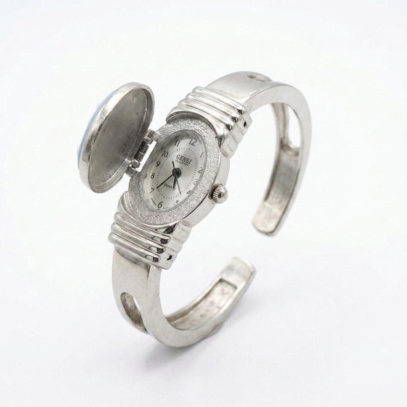 May include: A silver-toned cuff watch with an oval face and a hinged cover. The watch face is white with black numerals and hands. The bezel is encrusted with small, sparkling stones. The watch is open, revealing the time.