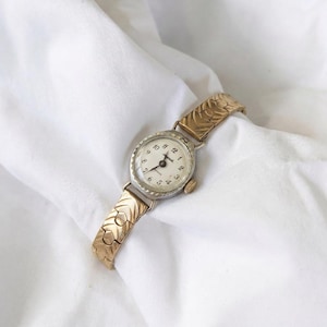 May include: A vintage wristwatch with a gold-toned band and a silver-toned face. The watch face has black numerals and hands against a white background. The band features a repeating leaf pattern. The watch is displayed on a white cloth.