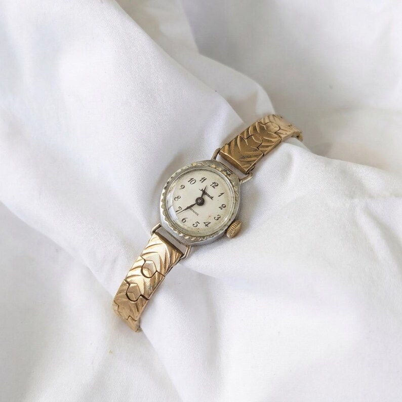 May include: A vintage wristwatch with a gold-toned band and a silver-toned face. The watch face has black numerals and hands against a white background. The band features a repeating leaf pattern. The watch is displayed on a white cloth.
