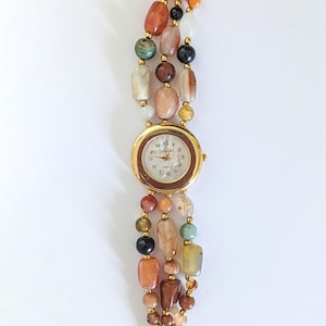 May include: A gold-toned wristwatch with a round face and a brown bezel. The watch band is made of three strands of colorful, irregularly shaped beads in shades of orange, brown, green, and cream.