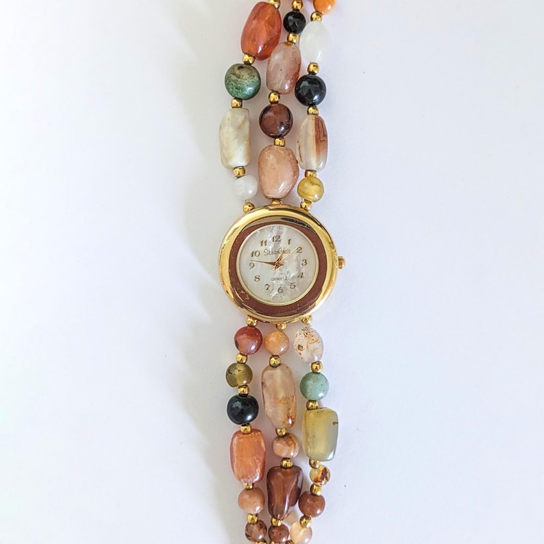 May include: A gold-toned wristwatch with a round face and a brown bezel. The watch band is made of three strands of colorful, irregularly shaped beads in shades of orange, brown, green, and cream.