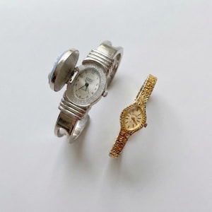 May include: Two vintage watches. One is a silver-toned hinged bangle watch with a light blue cameo and a white watch face with a diamond-like bezel. The other is a gold-toned watch with a textured band and a gold watch face.