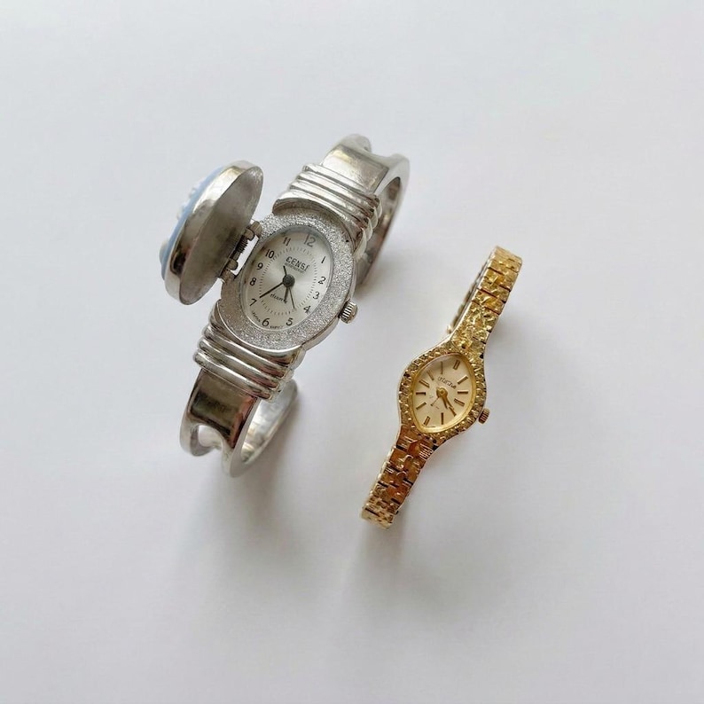May include: Two vintage watches. One is a silver-toned hinged bangle watch with a light blue cameo and a white watch face with a diamond-like bezel. The other is a gold-toned watch with a textured band and a gold watch face.