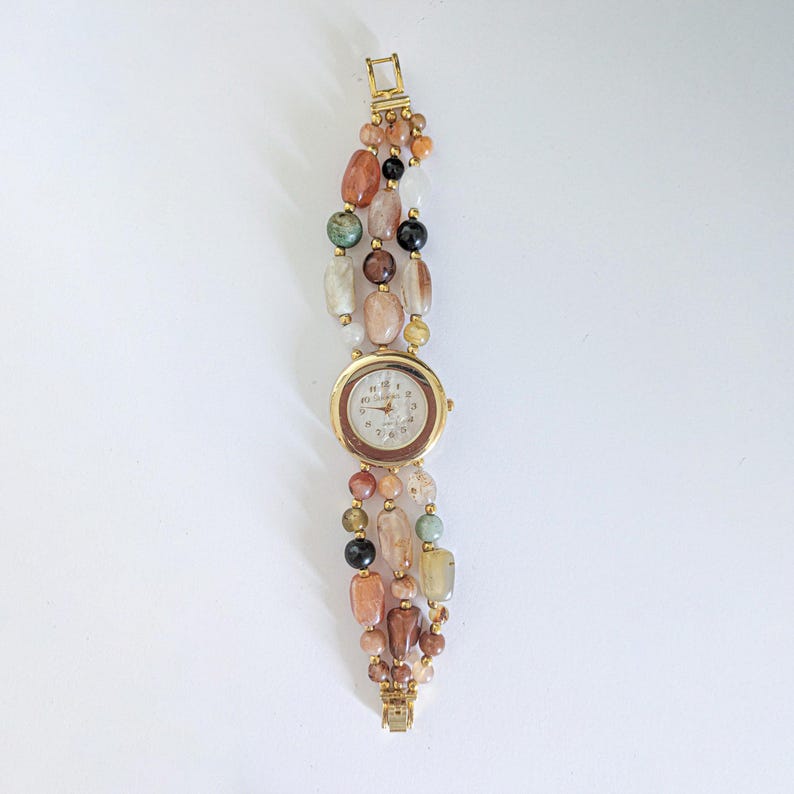 May include: A gold-toned wristwatch with a round face and a multi-strand beaded band. The band features a variety of colorful, irregularly shaped beads in shades of brown, beige, green, and black. The watch face has gold hands and hour markers.
