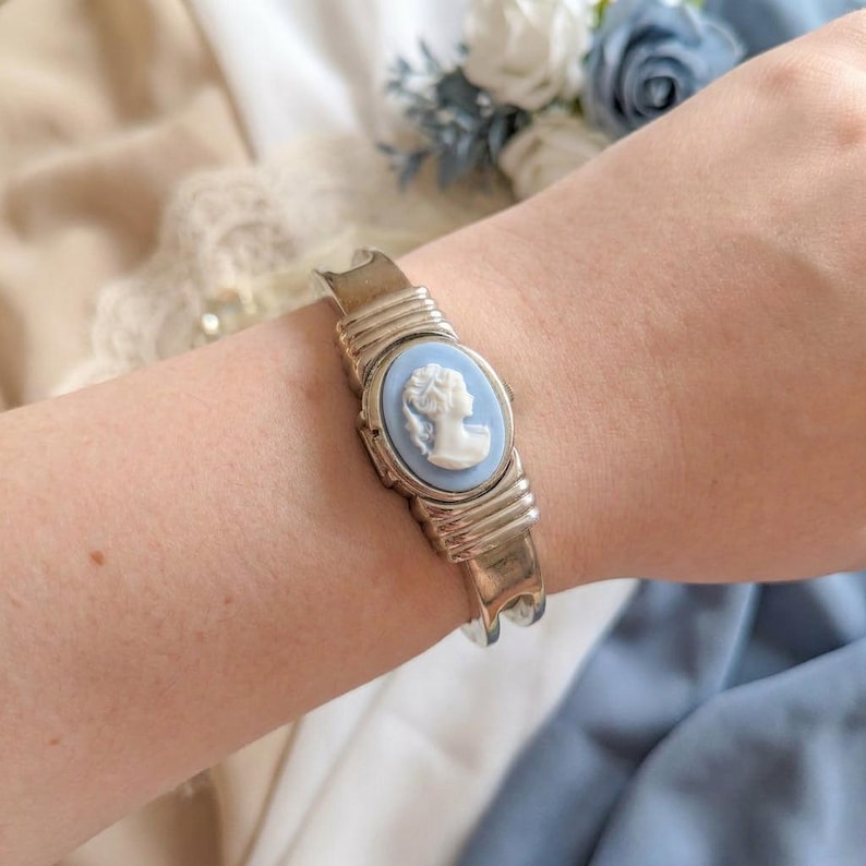 May include: Silver-toned metal cuff bracelet featuring a light blue cameo with a white profile. The oval cameo is set in a silver frame, and the bracelet has a textured design. A classic piece of jewelry.
