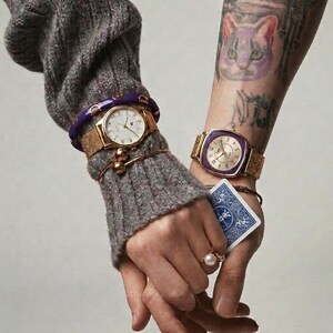 Peut inclure: Deux montres dorées avec des bracelets en maille et des cadrans blancs sont visibles. L'une a un cadran rond, l'autre un cadran carré avec une lunette violette. Une carte à jouer est tenue dans une main. Gros plan de deux poignets.