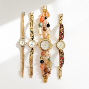 May include: Four gold-toned wristwatches with different band styles. One watch has a beaded band with various colors, while others have patterned or chain-link bands. Each watch has a round face with a light-colored dial.